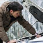 Man in a puffy winter jacket leaning over the open hood of a white vehicle with snowy trees in the background