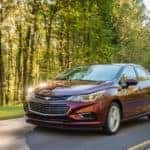 Maroon 2016 Chevrolet Cruze driving down a paved road past tall green trees