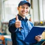 Mechanic in a blue jumpsuit and hat holding a clipboard and pen while giving a thumbs up
