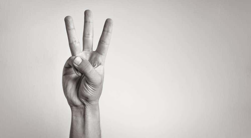Man in a blue sweater, in front of a grey wall, holding up three fingers