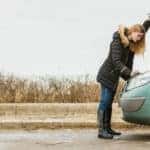 Woman in a black jacket looking under the hood of her blue vehicle that has broken down