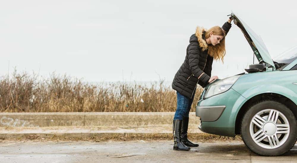 Woman in a black jacket looking under the hood of her blue vehicle that has broken down