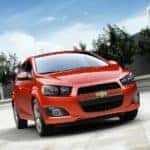 Orange 2016 Chevrolet Sonic parked in an empty lot with trees in the background