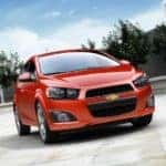 Orange 2016 Chevrolet Sonic parked on a tiled ground with trees and stone walls behind it