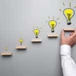 Five small wooden shelfs on a grey wall with yellow lightbulbs drawn above each one