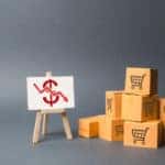Stack of cardboard boxes with shopping carts on them next to an easel with a money sign and downward arrow on it