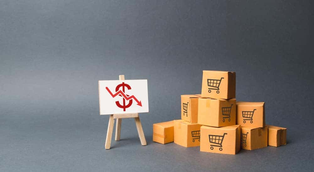 Stack of cardboard boxes with shopping carts on them next to an easel with a money sign and downward arrow on it