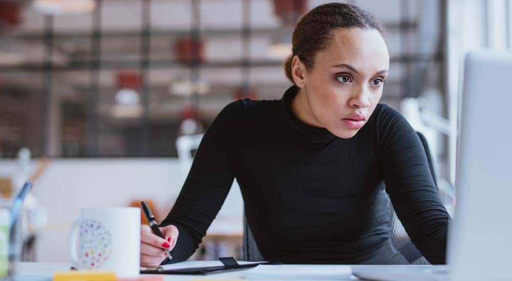 Woman in a black shirt looking intently at a white laptop while writing with a black pen