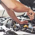 Man in a grey t-shirt and blue overalls using a wrench under the open hood of a vehicle