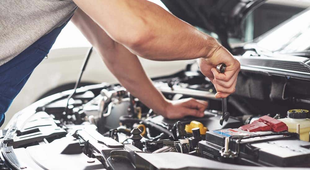 Man in a grey t-shirt and blue overalls using a wrench under the open hood of a vehicle
