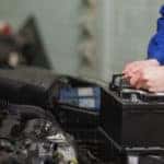 Man in a blue jumpsuit holding onto a car battery that is lifted out of a vehicle's open hood
