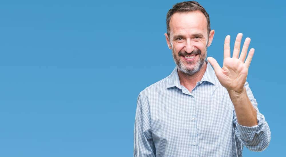 Man in a blue button-up shirt holding up five fingers in front of a blue wall