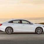 White 2016 Chevrolet Malibu parked on the side of a paved road in front of a body of water