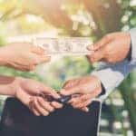 Two hands making a trade, one with a car key, the other with cash, in front of a white vehicle