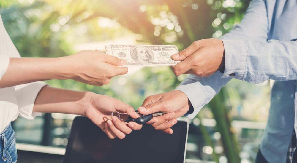 Two hands making a trade, one with a car key, the other with cash, in front of a white vehicle
