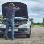 Man in blue jeans with his hands on his head in distress standing in front of the open hood of his broken-down vehicle