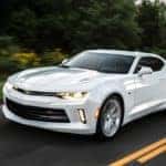 White 2016 Chevrolet Camaro performance vehicle driving on a paved road next to tall, dark green trees