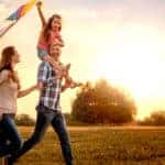 A mom and a dad with their daughter on his shoulders running through a field with a kite