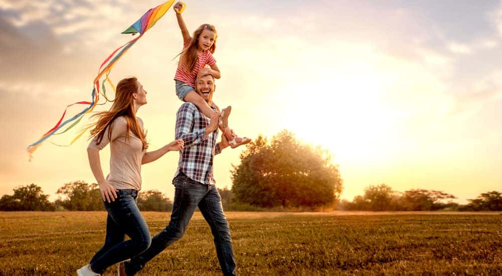 A mom and a dad with their daughter on his shoulders running through a field with a kite