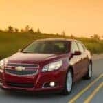 Red 2013 Chevrolet Malibu sedan driving down a paved road in front of an orange sky