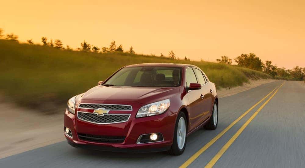 Red 2013 Chevrolet Malibu sedan driving down a paved road in front of an orange sky