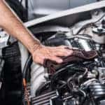 Hand wiping down the engine under the open hood of a vehicle with a brown cloth