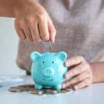 Hand placing change into a blue piggybank that is sitting on top of a pile of change