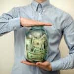 Man in a blue button-up shirt holding a large jar filled with cash