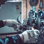 Mechanic in a black jacket and white gloves holding onto and inspecting an engine