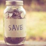 Mason jar full of coins with the word "SAVE" on it sitting on a brown ledge