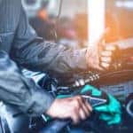 Mechanic in a blue shirt leaning over the open hood of a vehicle and giving a thumbs up