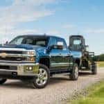 Deep blue 2017 Chevrolet Silverado pulling heavy machinery down a dirt road past green grass fields