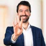 Man with facial hair in a suit holding three fingers up and smiling