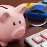 Pink piggybank, tan calculator, and a magnifying glass focused on a toy car all sitting on a brown surface