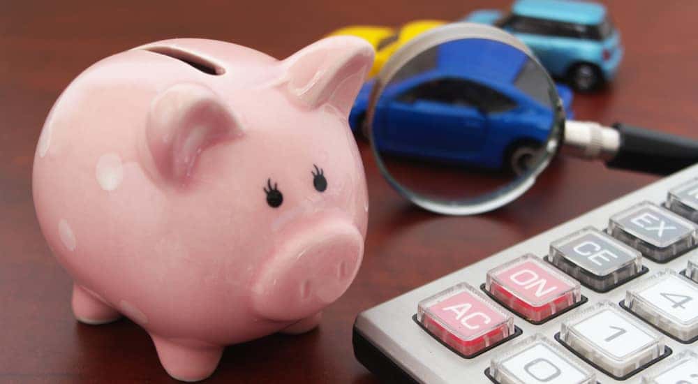 Pink piggybank, tan calculator, and a magnifying glass focused on a toy car all sitting on a brown surface