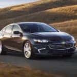 Black 2017 Chevrolet Malibu sedan driving down a paved path past brown, grassy hills
