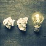 Four pieces of crumpled paper and a lit-up light bulb on a grey table