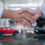 Men in button-up shirts shaking hands above a desk with a car key, stack of coins, and red toy car sitting on it