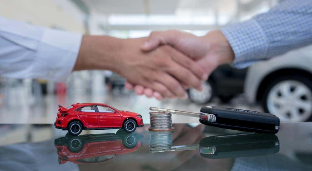Men in button-up shirts shaking hands above a desk with a car key, stack of coins, and red toy car sitting on it