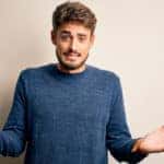 Man in a blue sweater shrugging in front of an off-white wall