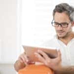 Man in a white button-up shirt sitting in an orange chair and reading on a silver tablet
