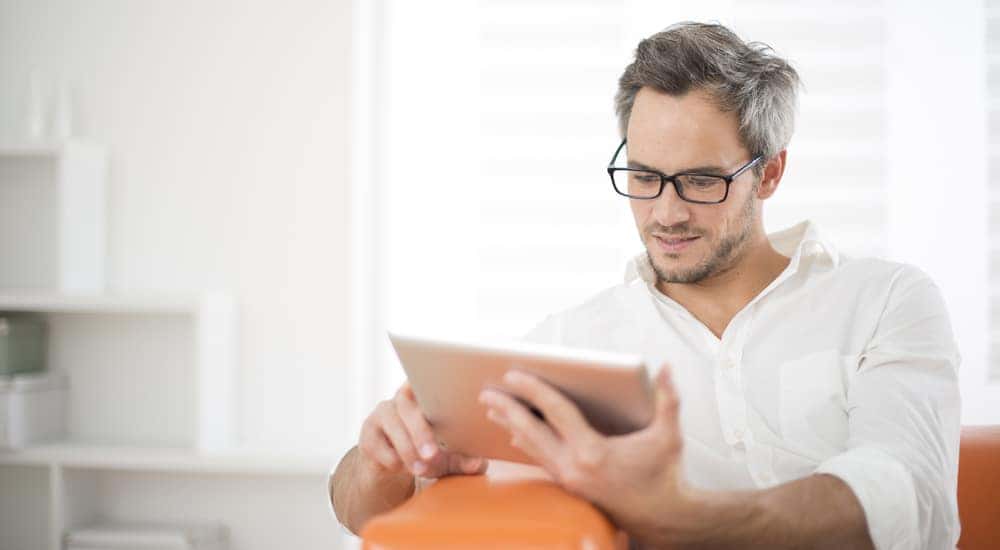 Man in a white button-up shirt sitting in an orange chair and reading on a silver tablet