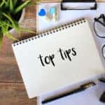 Notebook with the words "top tips" written on it sitting on top of a clipboard next to eyeglasses, a stamp, a pen, and a plant