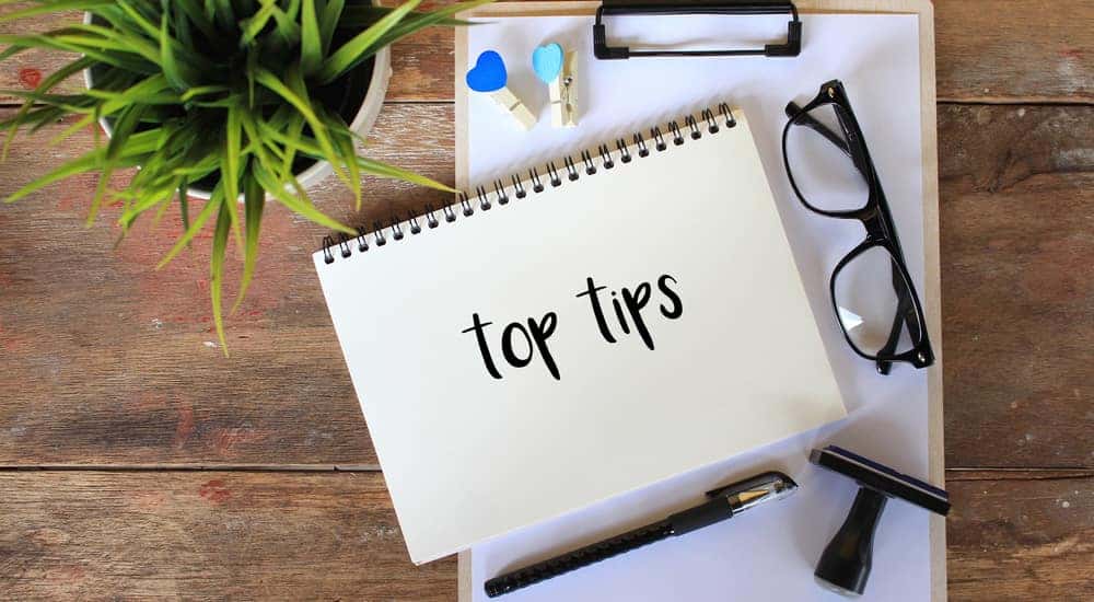 Notebook with the words "top tips" written on it sitting on top of a clipboard next to eyeglasses, a stamp, a pen, and a plant