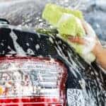 Hand holding a bright yellow sponge and scrubbing a soapy black vehicle