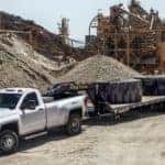 White 2017 Chevrolet Silverado 3500HD pulling a flat trailer with large tires on it through an all-dirt construction site