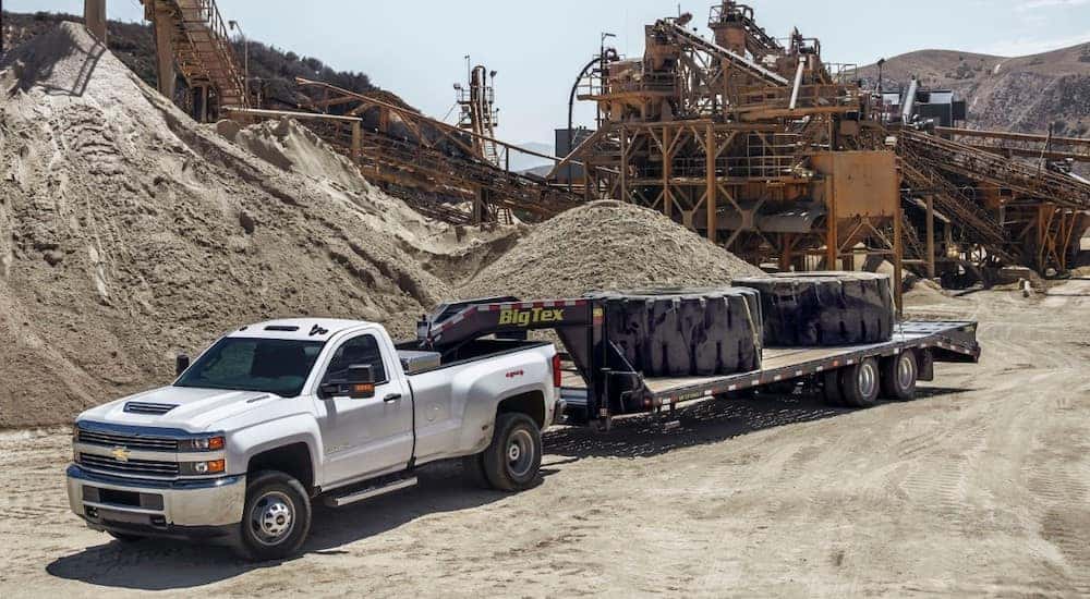 White 2017 Chevrolet Silverado 3500HD pulling a flat trailer with large tires on it through an all-dirt construction site