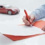 Hand holding a red pen and signing a car loan on a desk next to a red toy car