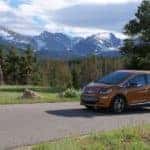 Orange 2017 Chevrolet Bolt EV driving on a paved road past grass, trees, and mountains