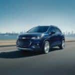 Dark blue 2018 Chevrolet Trax driving down a paved road past a body of water and a city skyline